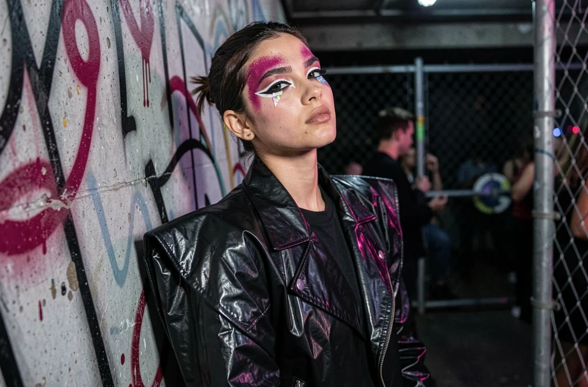 Modèle non-binaire arborant un maquillage club kid avec liner blanc graphique et pigments holographiques dans un décor de club underground