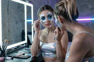 Jeune femme appliquant un maquillage techno pouffe avec des pigments iridescents et du glitter devant un miroir à LED